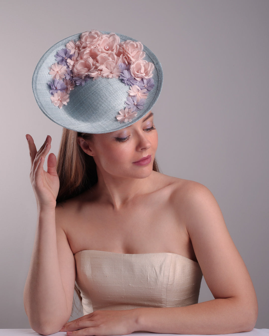 The model is showcasing the front top angle of a blue classic saucer hat with curved hight on the back left worn towards the right side of the head. The top of the hat is decorated with pink and purple flower&amp;nbsp;arrangement. The hat is fitted with a wire headband and an elastic.