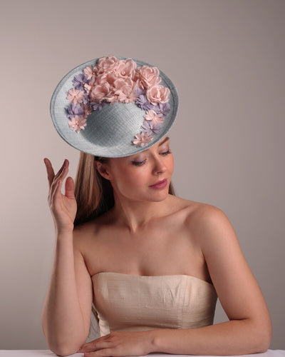 The model is showcasing the front top angle of a blue classic saucer hat with curved hight on the back left worn towards the right side of the head. The top of the hat is decorated with pink and purple flower&nbsp;arrangement. The hat is fitted with a wire headband and an elastic.