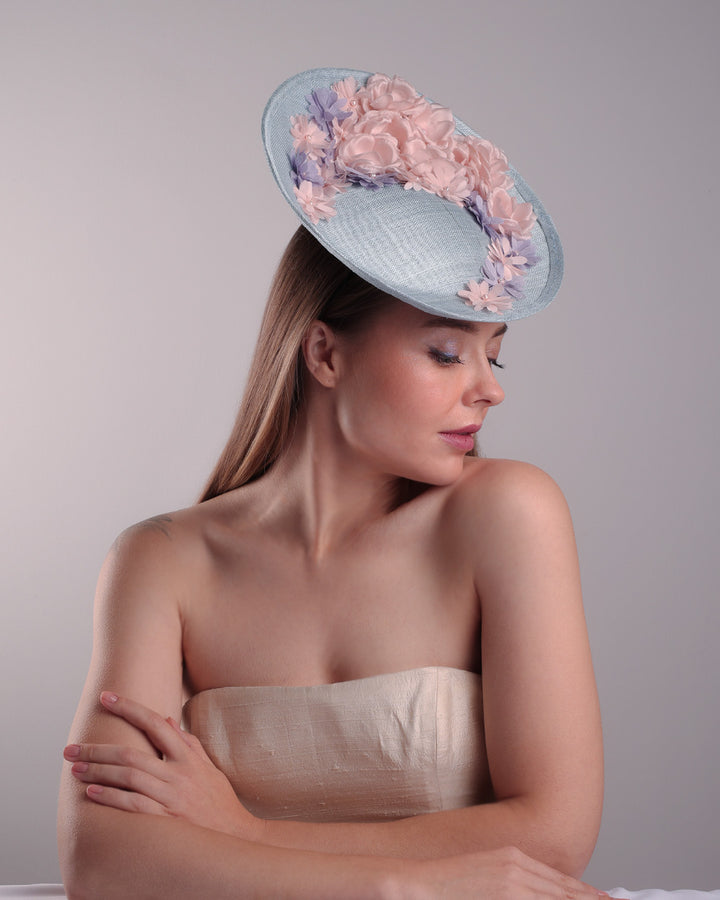 The model is showcasing the right angle of a blue classic saucer hat with curved hight on the back left worn towards the right side of the head. The top of the hat is decorated with pink and purple flower&amp;nbsp;arrangement. The hat is fitted with a wire headband and an elastic.
