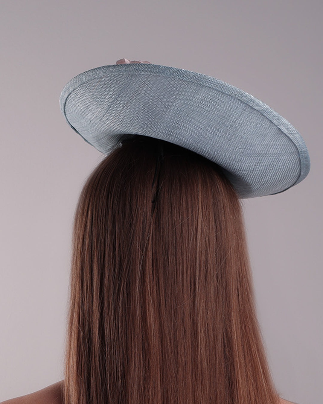 The model is showcasing the back of a blue classic saucer hat with curved hight on the back left worn towards the right side of the head. The top of the hat is decorated with pink and purple flower&amp;nbsp;arrangement. The hat is fitted with a wire headband and an elastic.