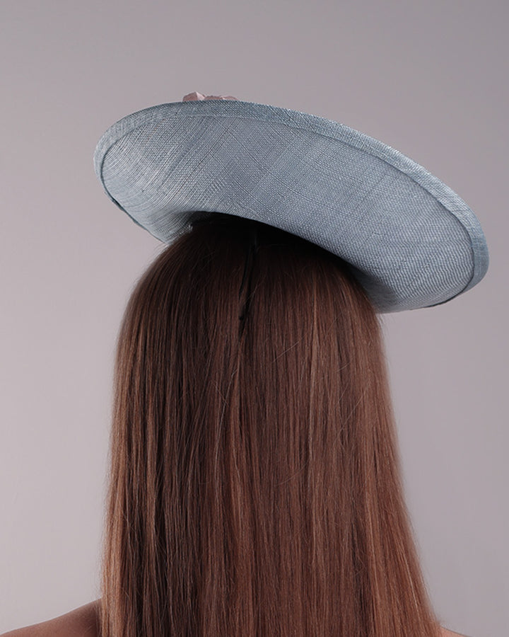 The model is showcasing the back of a blue classic saucer hat with curved hight on the back left worn towards the right side of the head. The top of the hat is decorated with pink and purple flower&amp;nbsp;arrangement. The hat is fitted with a wire headband and an elastic.