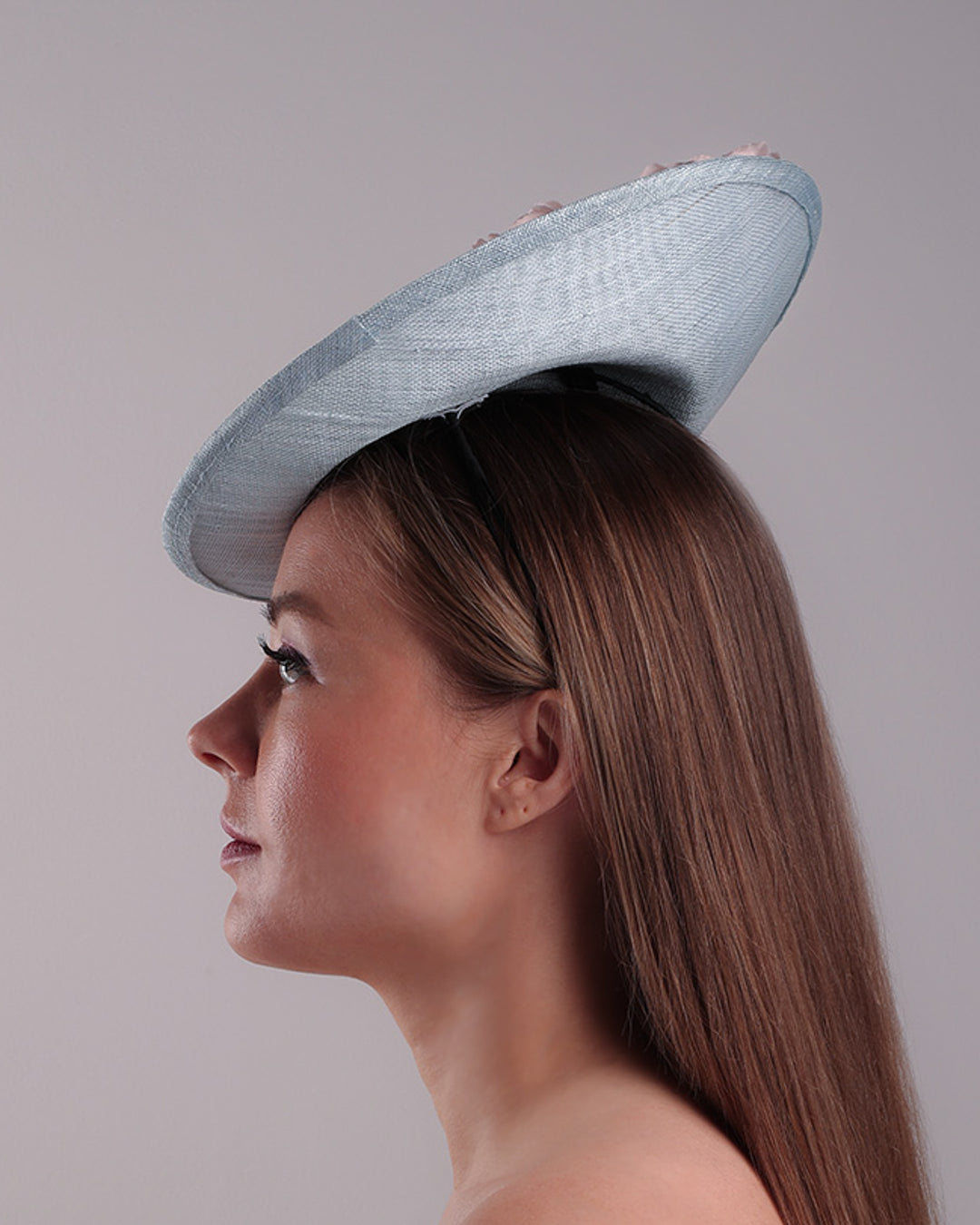 The model is showcasing the left side of a blue classic saucer hat with curved hight on the back left worn towards the right side of the head. The top of the hat is decorated with pink and purple flower&amp;nbsp;arrangement. The hat is fitted with a wire headband and an elastic.