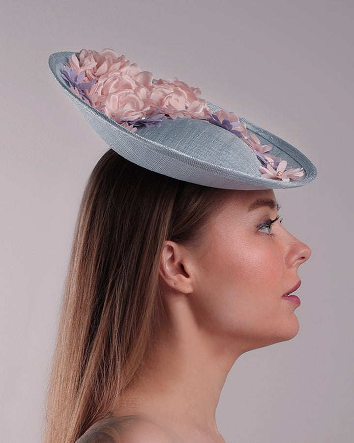 The model is showcasing the right side of a blue classic saucer hat with curved hight on the back left worn towards the right side of the head. The top of the hat is decorated with pink and purple flower&amp;nbsp;arrangement. The hat is fitted with a wire headband and an elastic.