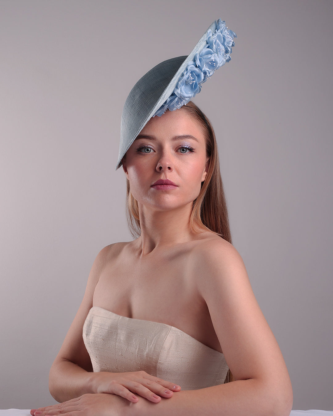Model is showcasing the front of the blue saucer hat with an high angle decorated with blue silk flowers in the left high inner side, the hat is fitted on a mini saucer that is secured to the head with &amp;nbsp;a wire headband and elastic as well as a comb.