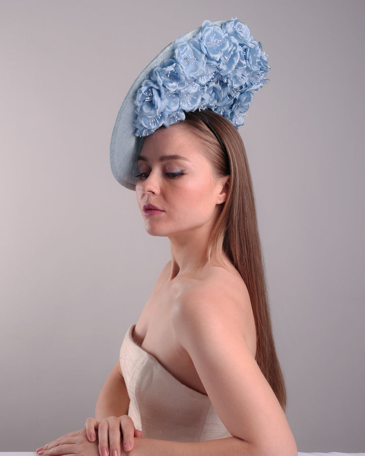Model is showcasing the front left side of the blue saucer hat with an high angle decorated with blue silk flowers in the left high inner side, the hat is fitted on a mini saucer that is secured to the head with;a wire headband and elastic as well as a comb.
