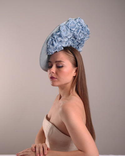 Model is showcasing the front left side of the blue saucer hat with an high angle decorated with blue silk flowers in the left high inner side, the hat is fitted on a mini saucer that is secured to the head with &nbsp;a wire headband and elastic as well as a comb.