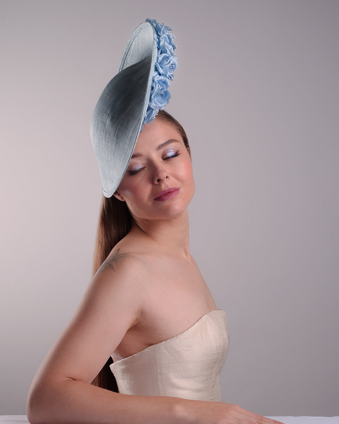 Model is showcasing the front right side of the blue saucer hat with an high angle decorated with blue silk flowers in the left high inner side, the hat is fitted on a mini saucer that is secured to the head with &amp;nbsp;a wire headband and elastic as well as a comb.