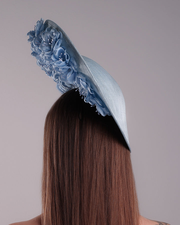 Model is showcasing the back of the blue saucer hat with an high angle decorated with blue silk flowers in the left high inner side, the hat is fitted on a mini saucer that is secured to the head with &amp;nbsp;a wire headband and elastic as well as a comb.