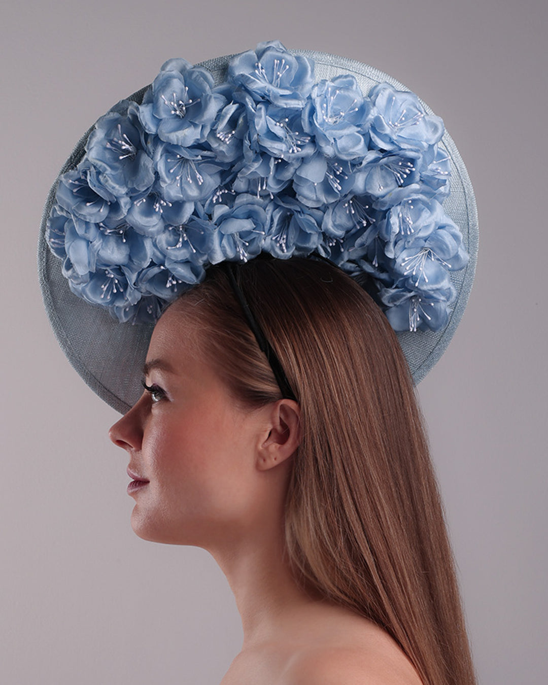 Model is showcasing the left side of the blue saucer hat with an high angle decorated with blue silk flowers in the left high inner side, the hat is fitted on a mini saucer that is secured to the head with &amp;nbsp;a wire headband and elastic as well as a comb.