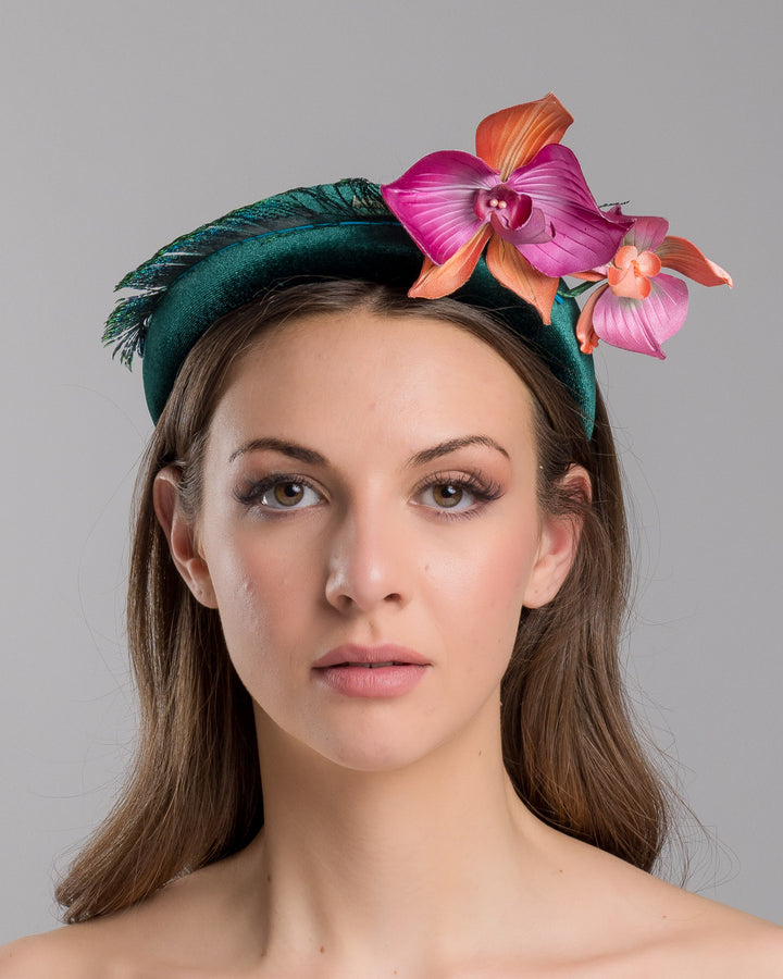 The model is showcasing the front side close up of the emerald green velvet padded headband embellished with pink and peach orchid flowers and a delicate fan of dark green feather trim along one side. Designed and made by Merve Bayindir Millinery.
