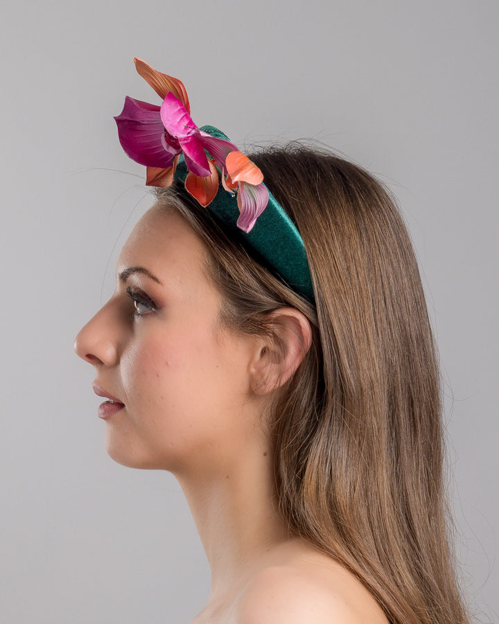 The model is showcasing the left side close up of the emerald green velvet padded headband embellished with pink and peach orchid flowers and a delicate fan of dark green feather trim along one side. Designed and made by Merve Bayindir Millinery.

