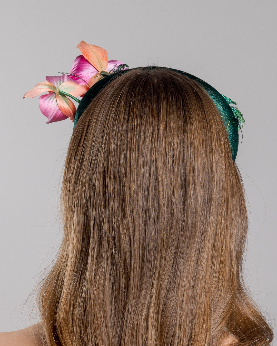 The model is showcasing the back side close up of the emerald green velvet padded headband embellished with pink and peach orchid flowers and a delicate fan of dark green feather trim along one side. Designed and made by Merve Bayindir Millinery.

