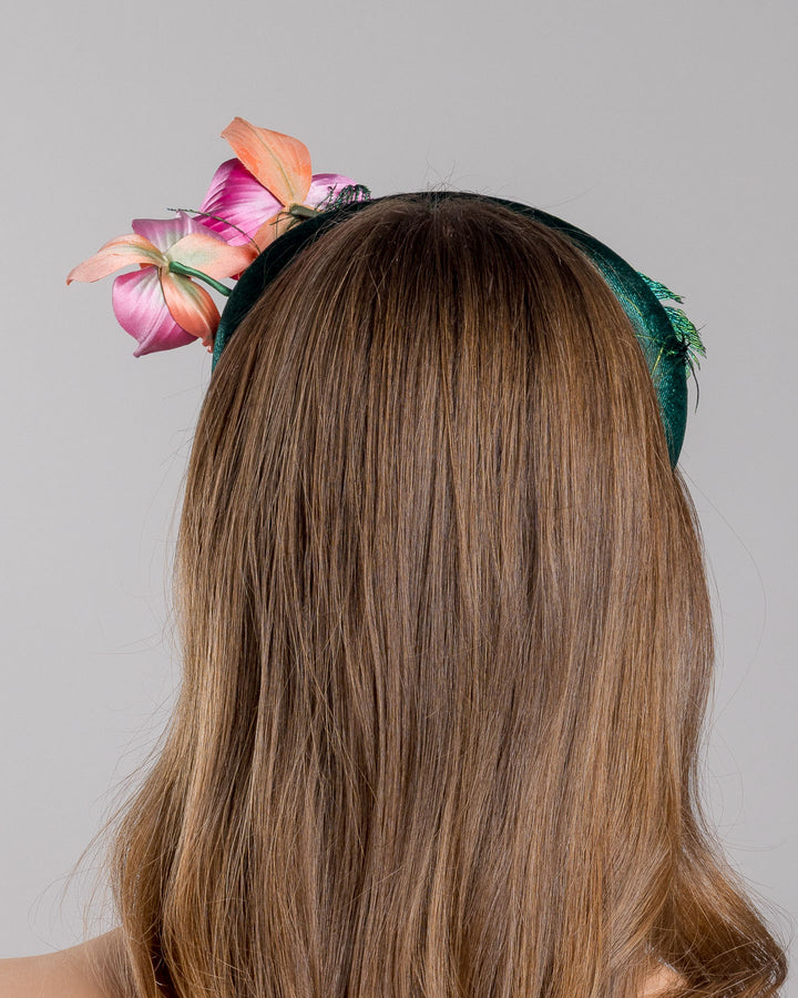 The model is showcasing the back side close up of the emerald green velvet padded headband embellished with pink and peach orchid flowers and a delicate fan of dark green feather trim along one side. Designed and made by Merve Bayindir Millinery.
