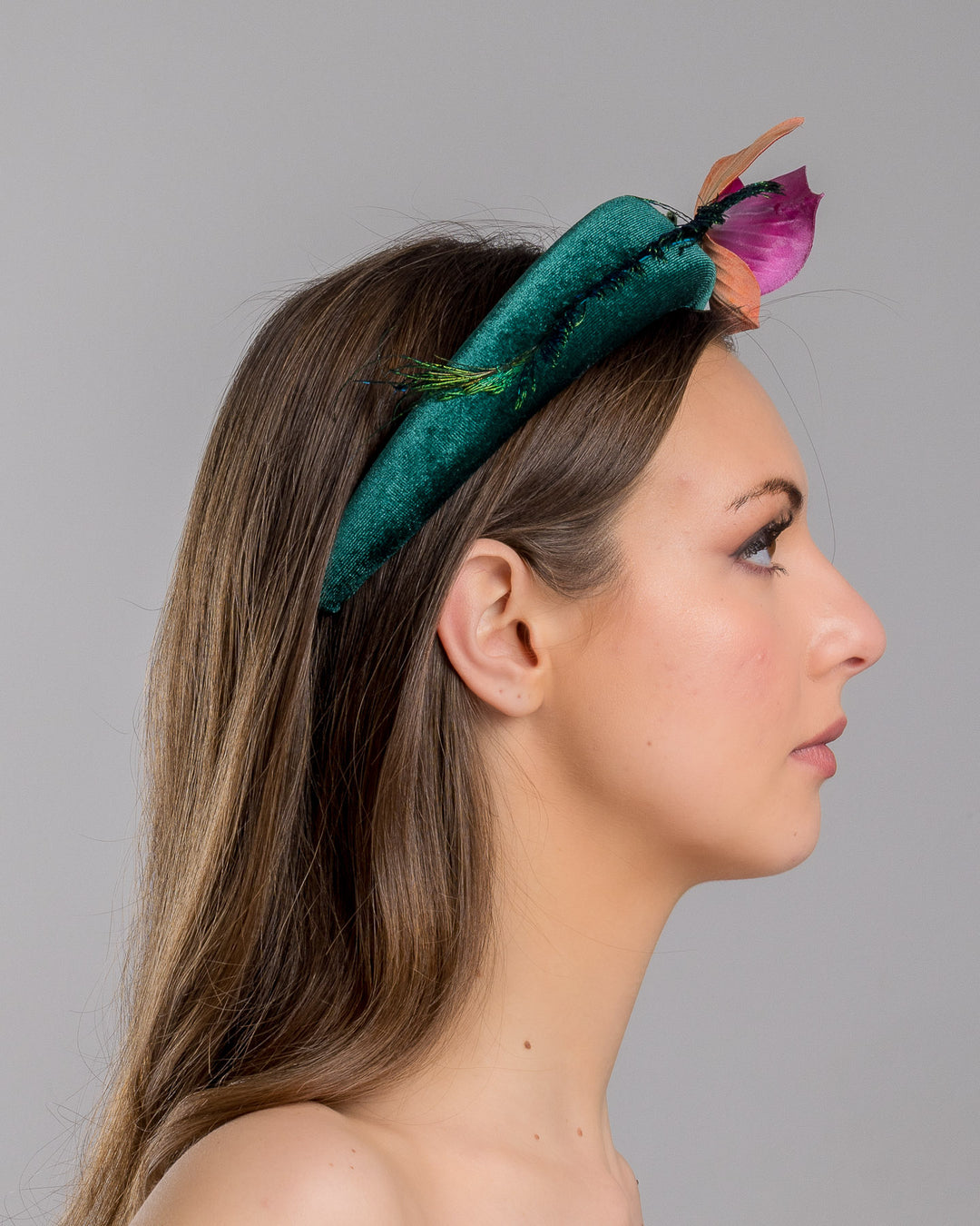 The model is showcasing the right side close up of the emerald green velvet padded headband embellished with pink and peach orchid flowers and a delicate fan of dark green feather trim along one side. Designed and made by Merve Bayindir Millinery.

