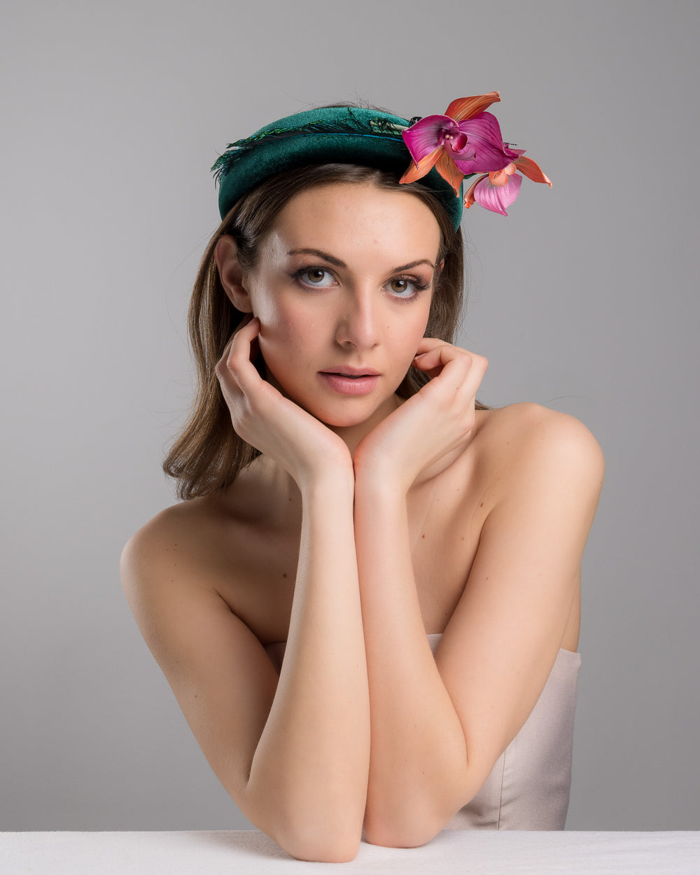 The female model is posing, wearing
Ballet, the emerald green velvet padded headband embellished with pink and peach orchid flowers and a delicate fan of dark green feather trim along one side. Designed and made by Merve Bayindir Millinery.
