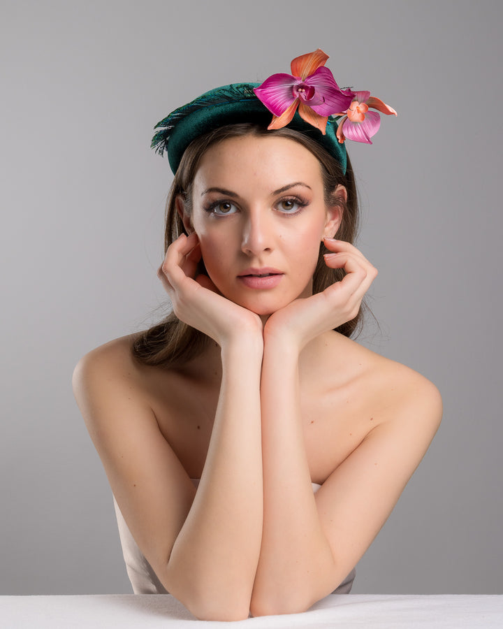 The female model is posing, wearing
Ballet, the emerald green velvet padded headband embellished with pink and peach orchid flowers and a delicate fan of dark green feather trim along one side. Designed and made by Merve Bayindir Millinery.
