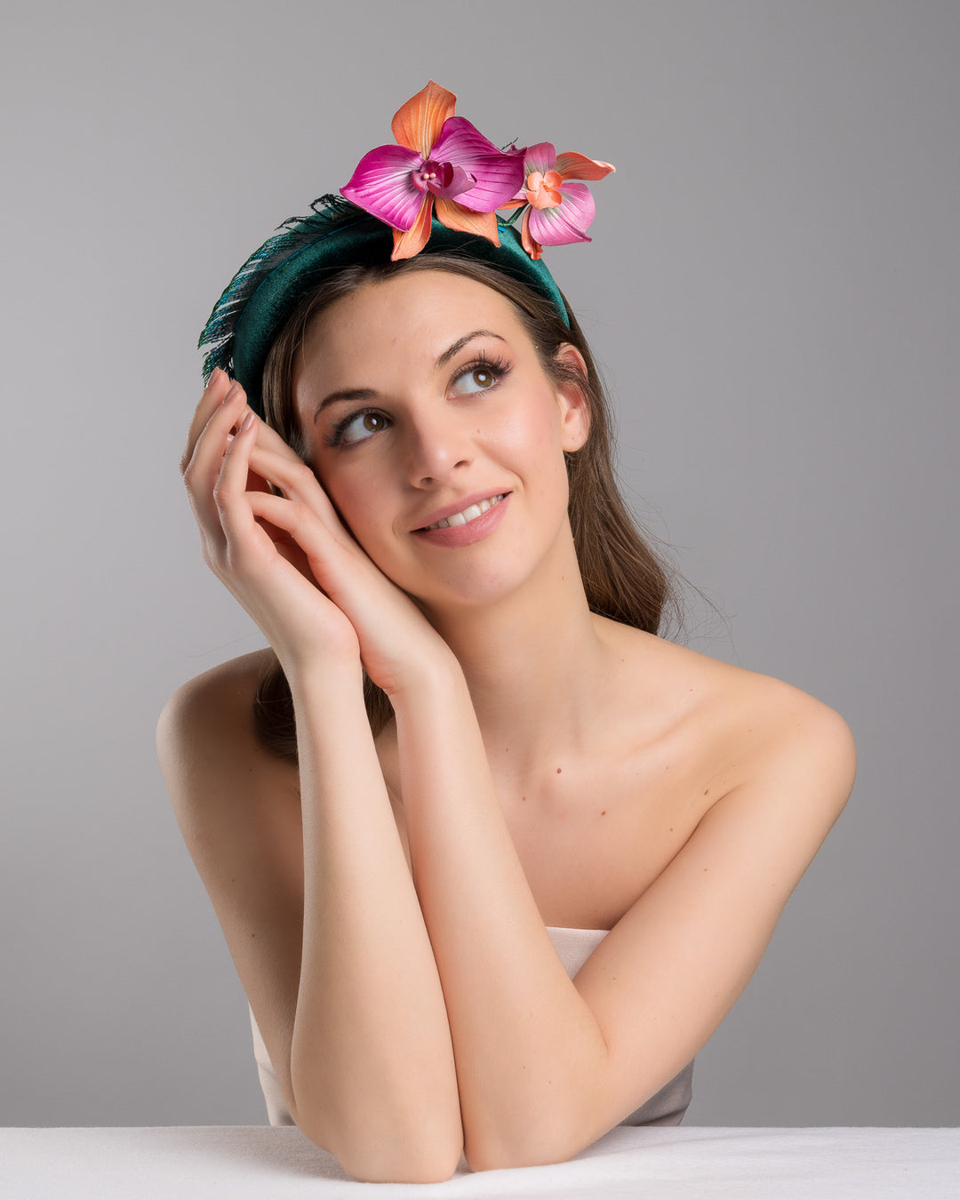 The female model is posing, wearing
Ballet, the emerald green velvet padded headband embellished with pink and peach orchid flowers and a delicate fan of dark green feather trim along one side. Designed and made by Merve Bayindir Millinery.
