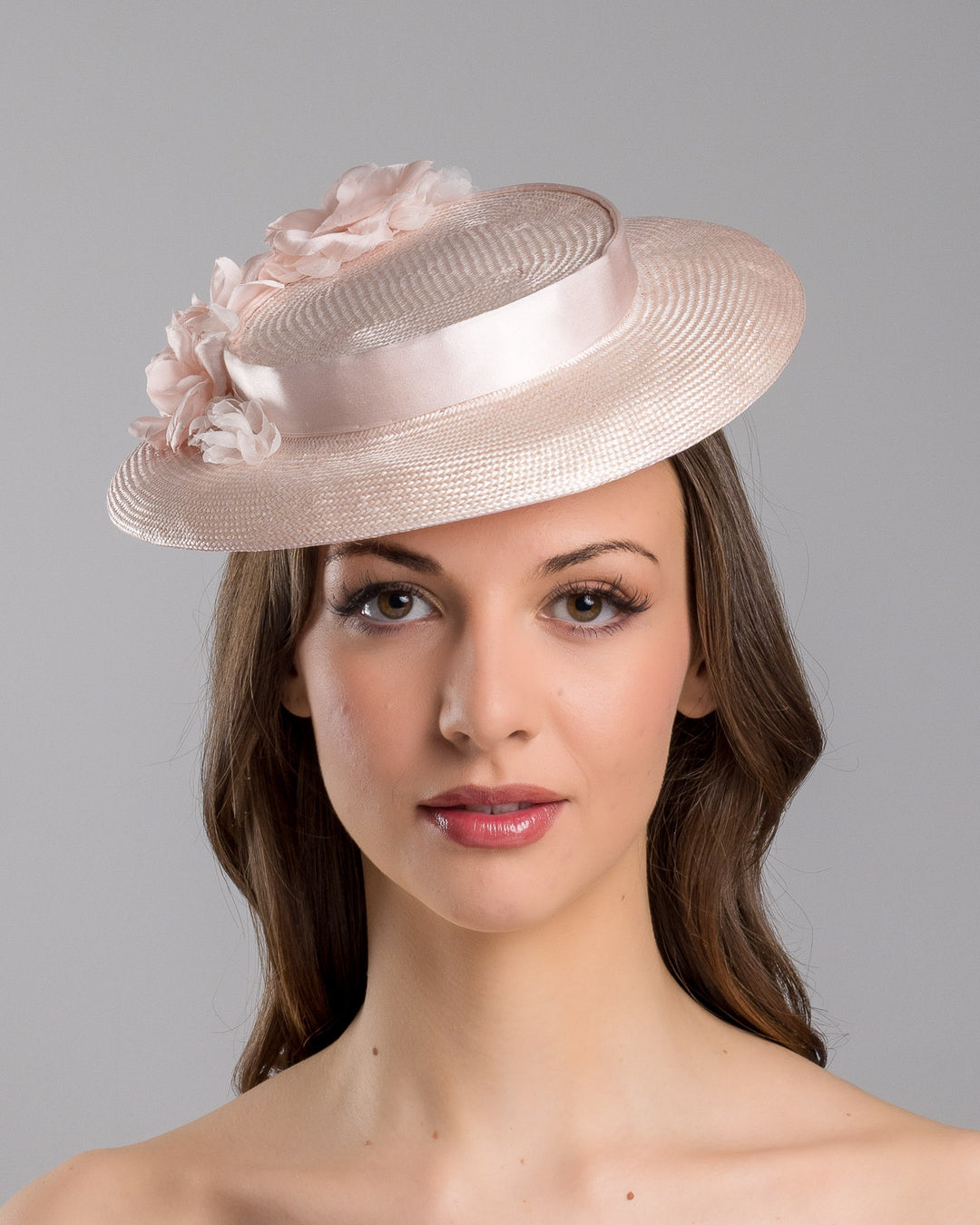 The model is showcasing the front side close up of the blush pink mini boater hat crafted from woven straw, trimmed with a satin band and adorned with soft pink floral accents and a flowing satin ribbon at the back, mounted on a slim headband. Designed and made by Merve Bayindir Millinery.
