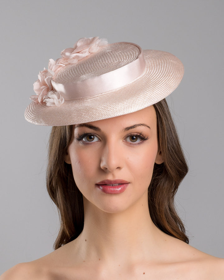 The model is showcasing the front side close up of the blush pink mini boater hat crafted from woven straw, trimmed with a satin band and adorned with soft pink floral accents and a flowing satin ribbon at the back, mounted on a slim headband. Designed and made by Merve Bayindir Millinery.
