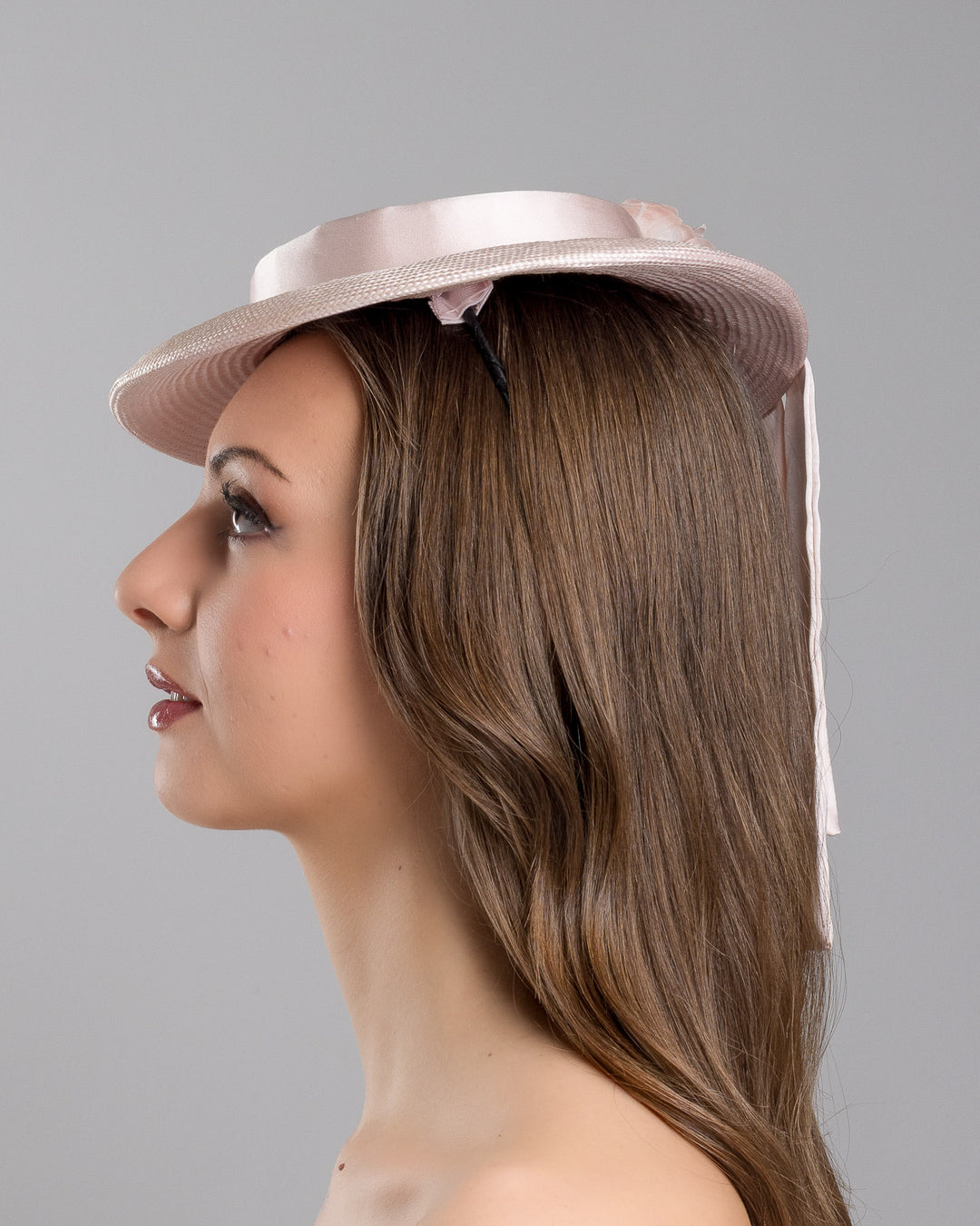 The model is showcasing the left side close up of the blush pink mini boater hat crafted from woven straw, trimmed with a satin band and adorned with soft pink floral accents and a flowing satin ribbon at the back, mounted on a slim headband. Designed and made by Merve Bayindir Millinery.
