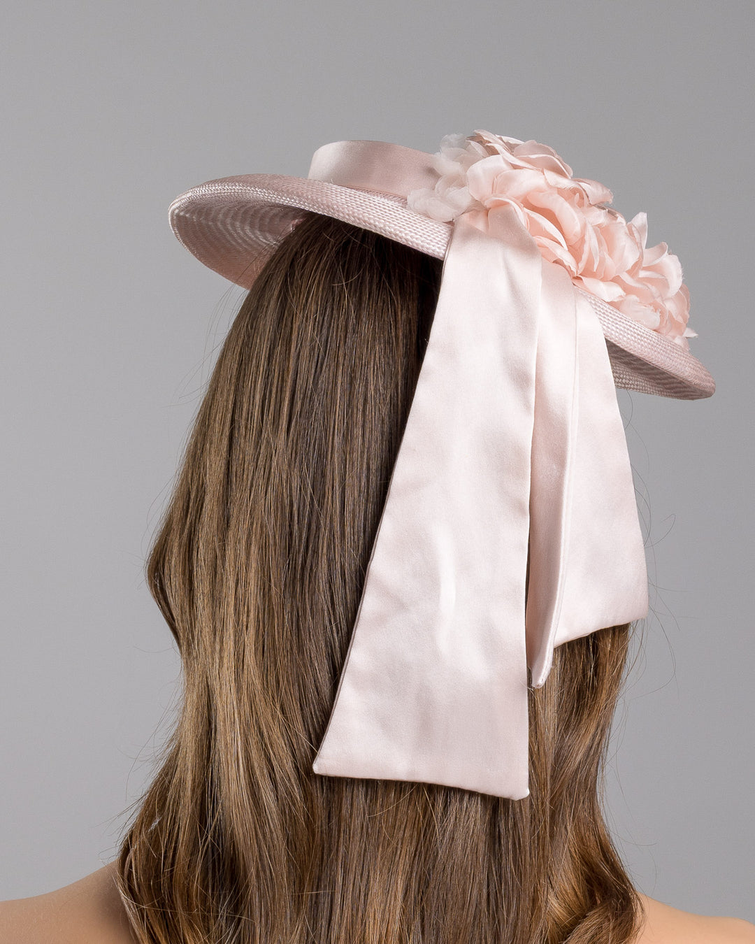 The model is showcasing the back side close up of the blush pink mini boater hat crafted from woven straw, trimmed with a satin band and adorned with soft pink floral accents and a flowing satin ribbon at the back, mounted on a slim headband. Designed and made by Merve Bayindir Millinery.
