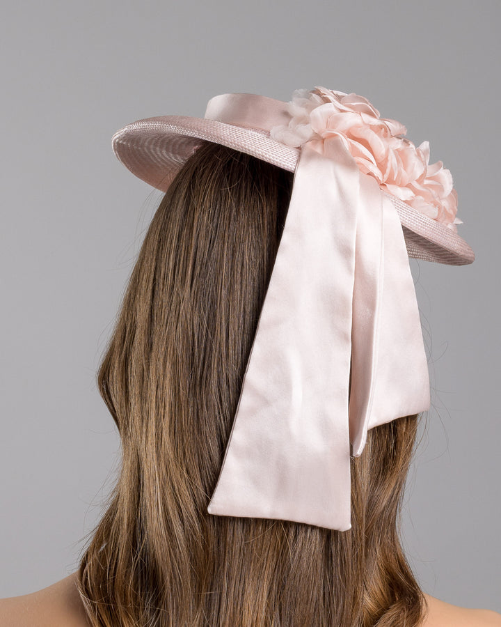 The model is showcasing the back side close up of the blush pink mini boater hat crafted from woven straw, trimmed with a satin band and adorned with soft pink floral accents and a flowing satin ribbon at the back, mounted on a slim headband. Designed and made by Merve Bayindir Millinery.
