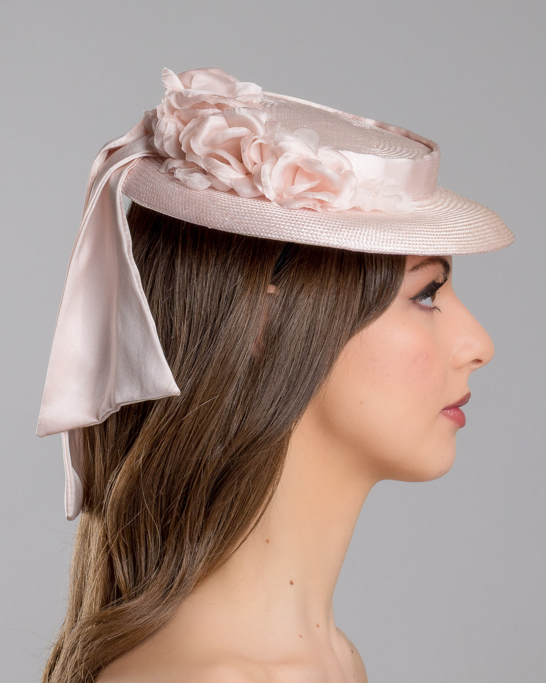 The model is showcasing the right side close up of the blush pink mini boater hat crafted from woven straw, trimmed with a satin band and adorned with soft pink floral accents and a flowing satin ribbon at the back, mounted on a slim headband. Designed and made by Merve Bayindir Millinery.
