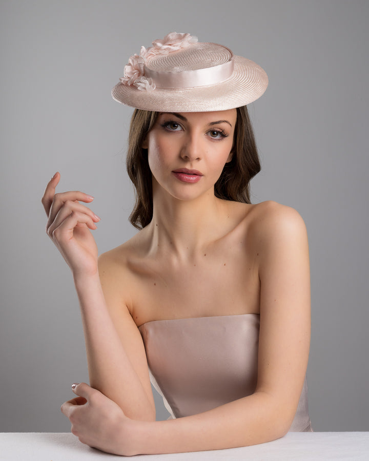 The female model is posing, wearing
Bellamy, the blush pink mini boater hat crafted from woven straw, trimmed with a satin band and adorned with soft pink floral accents and a flowing satin ribbon at the back, mounted on a slim headband. Designed and made by Merve Bayindir Millinery.
