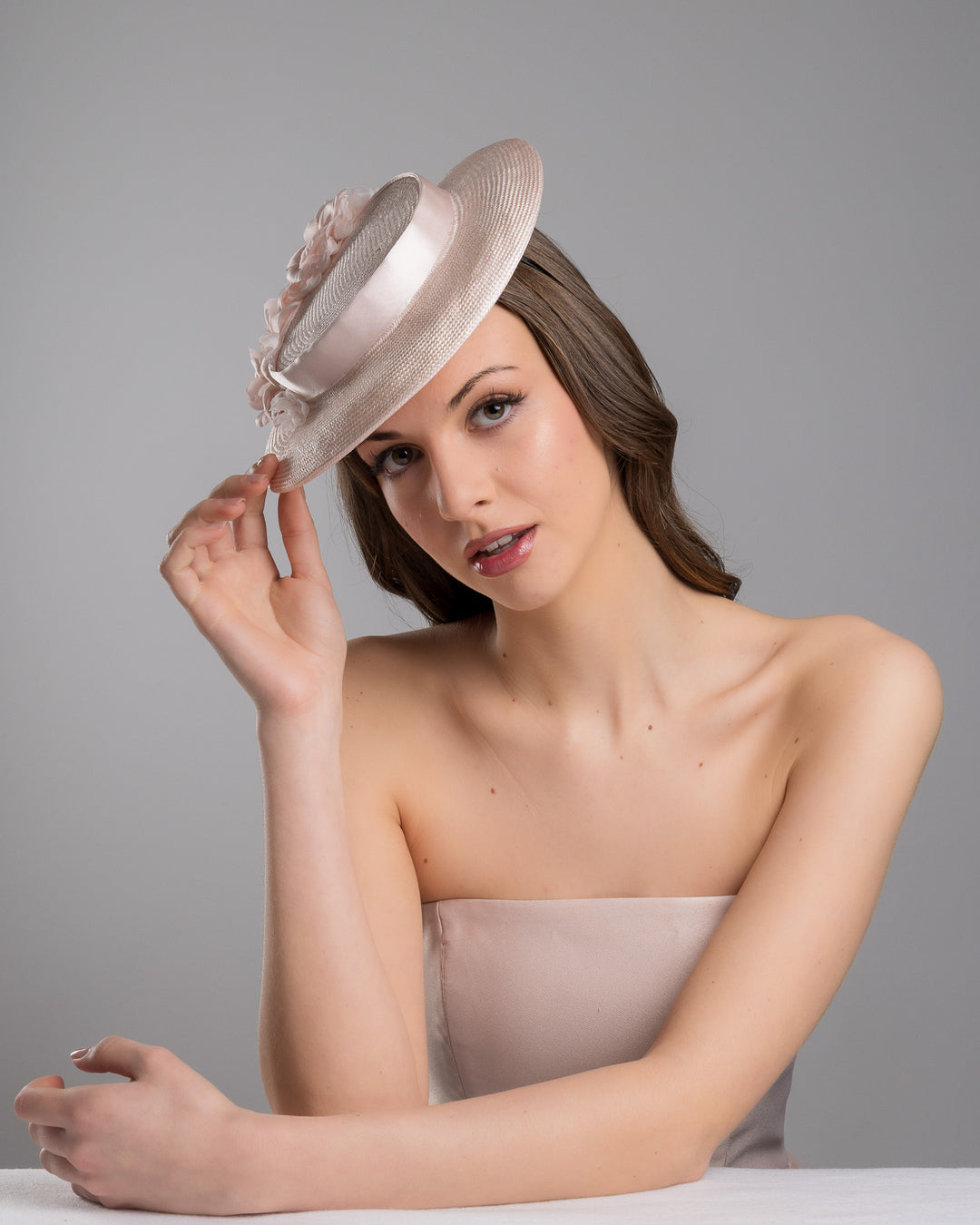 The female model is posing, wearing
Bellamy, the blush pink mini boater hat crafted from woven straw, trimmed with a satin band and adorned with soft pink floral accents and a flowing satin ribbon at the back, mounted on a slim headband. Designed and made by Merve Bayindir Millinery.
