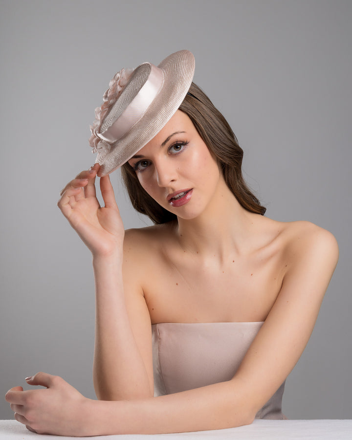 The female model is posing, wearing
Bellamy, the blush pink mini boater hat crafted from woven straw, trimmed with a satin band and adorned with soft pink floral accents and a flowing satin ribbon at the back, mounted on a slim headband. Designed and made by Merve Bayindir Millinery.
