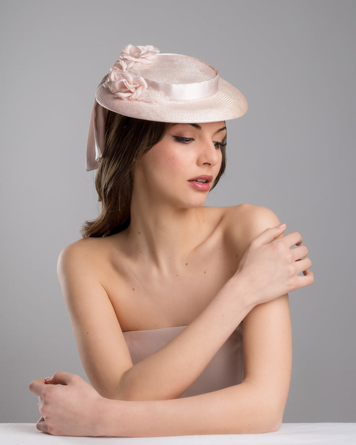 The female model is posing, wearing
Bellamy, the blush pink mini boater hat crafted from woven straw, trimmed with a satin band and adorned with soft pink floral accents and a flowing satin ribbon at the back, mounted on a slim headband. Designed and made by Merve Bayindir Millinery.
