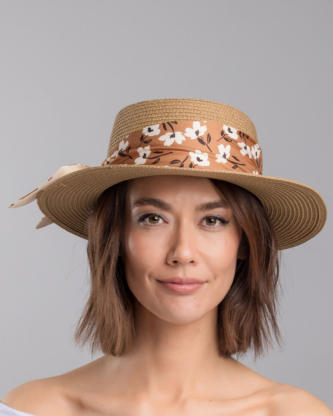 The model is showcasing the front side close up of the natural woven straw boater hat with a wide brim, adorned with a brown floral-print ribbon tied in a bow and finished with a small embroidered flower detail at the center. Designed and made by Merve Bayindir millinery.
