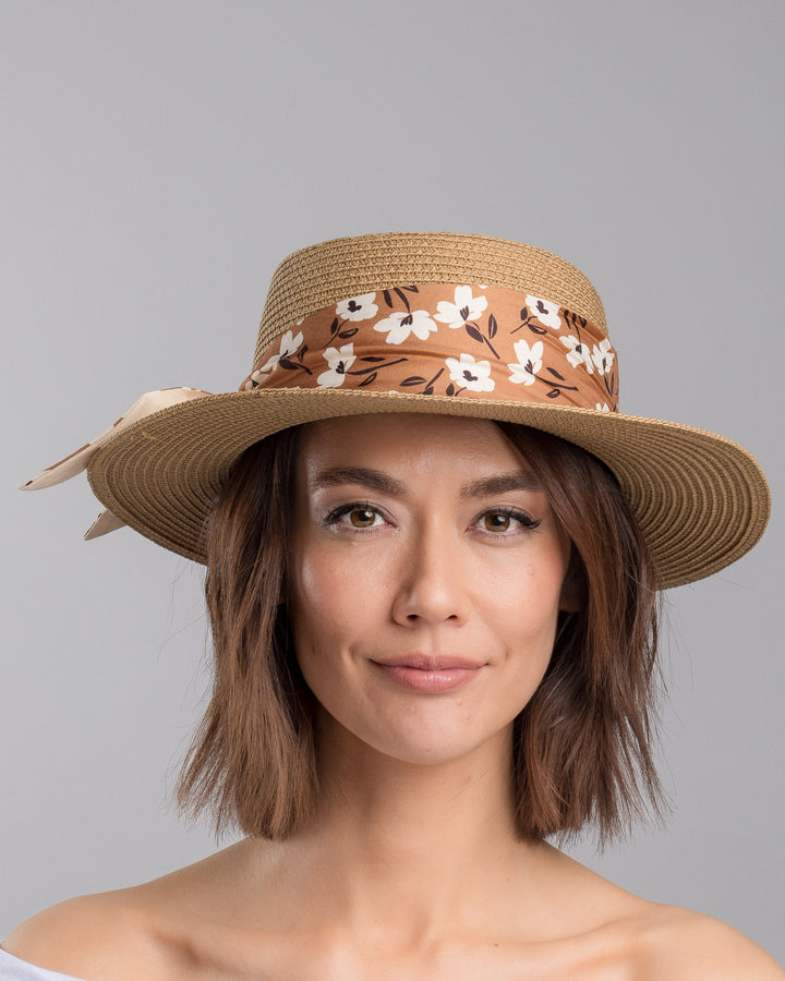 The model is showcasing the front side close up of the natural woven straw boater hat with a wide brim, adorned with a brown floral-print ribbon tied in a bow and finished with a small embroidered flower detail at the center. Designed and made by Merve Bayindir millinery.
