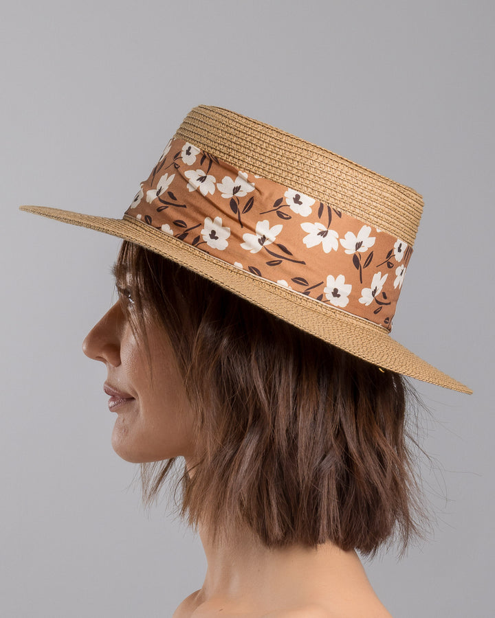 The model is showcasing the left side close up of the natural woven straw boater hat with a wide brim, adorned with a brown floral-print ribbon tied in a bow and finished with a small embroidered flower detail at the center. Designed and made by Merve Bayindir millinery.