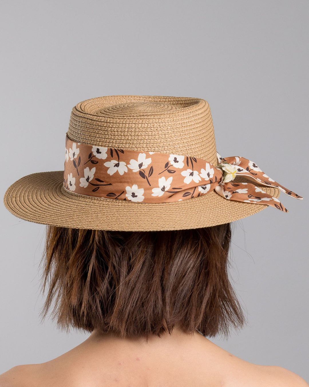 The model is showcasing the back side close up of the natural woven straw boater hat with a wide brim, adorned with a brown floral-print ribbon tied in a bow and finished with a small embroidered flower detail at the center. Designed and made by Merve Bayindir millinery.