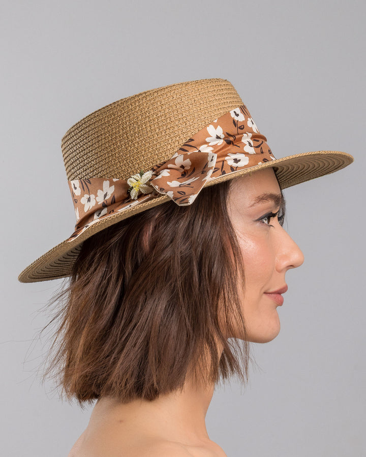 The model is showcasing the right side close up of the natural woven straw boater hat with a wide brim, adorned with a brown floral-print ribbon tied in a bow and finished with a small embroidered flower detail at the center. Designed and made by Merve Bayindir millinery.