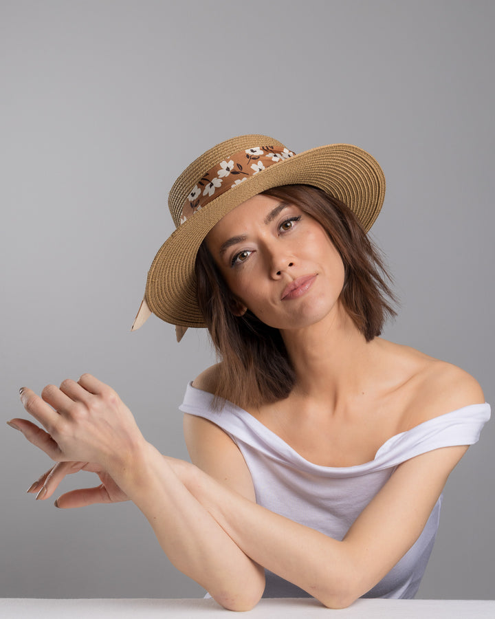 The female model is posing, wearing Brisa,  the natural woven straw boater hat with a wide brim, adorned with a brown floral-print ribbon tied in a bow and finished with a small embroidered flower detail at the center. Designed and made by Merve Bayindir millinery.