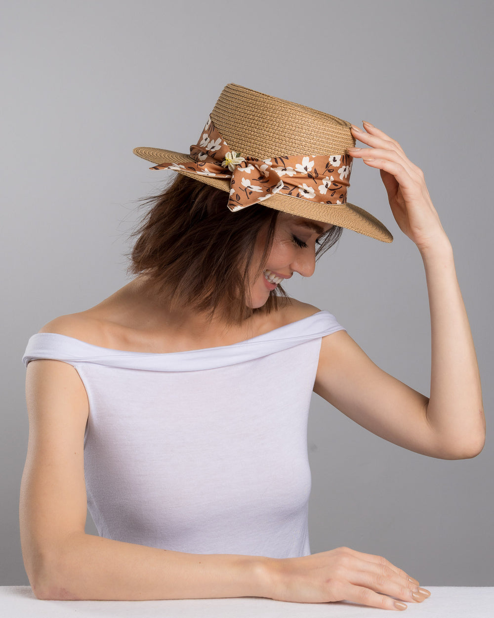 The female model is posing, wearing Brisa,  the natural woven straw boater hat with a wide brim, adorned with a brown floral-print ribbon tied in a bow and finished with a small embroidered flower detail at the center. Designed and made by Merve Bayindir millinery.