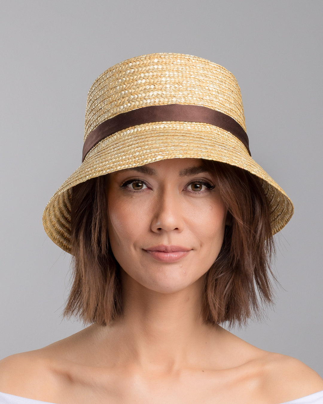 The model is showcasing the front side close up of the natural woven straw bucket hat with a softly sloping brim, finished with a brown ribbon band and long trailing ties at the side. Designed and Made by Merve Bayindir millinery. Designed and made by Merve Bayindir millinery.