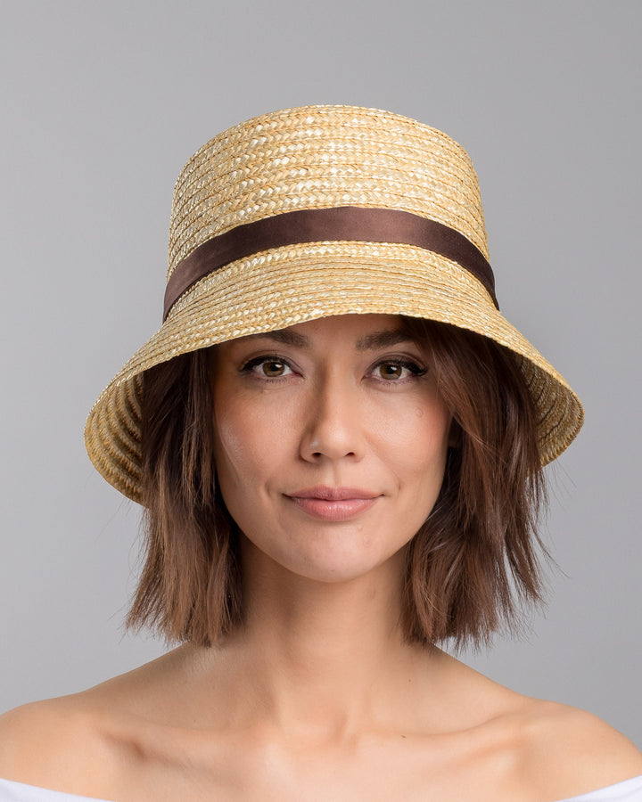The model is showcasing the front side close up of the natural woven straw bucket hat with a softly sloping brim, finished with a brown ribbon band and long trailing ties at the side. Designed and Made by Merve Bayindir millinery. Designed and made by Merve Bayindir millinery.