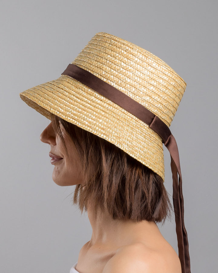 The model is showcasing the left side close up of the natural woven straw bucket hat with a softly sloping brim, finished with a brown ribbon band and long trailing ties at the side. Designed and Made by Merve Bayindir millinery. Designed and made by Merve Bayindir millinery.
