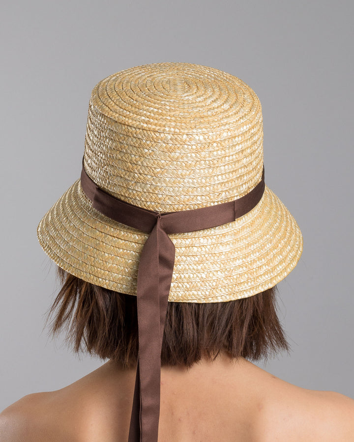 The model is showcasing the back side close up of the natural woven straw bucket hat with a softly sloping brim, finished with a brown ribbon band and long trailing ties at the side. Designed and Made by Merve Bayindir millinery. Designed and made by Merve Bayindir millinery.