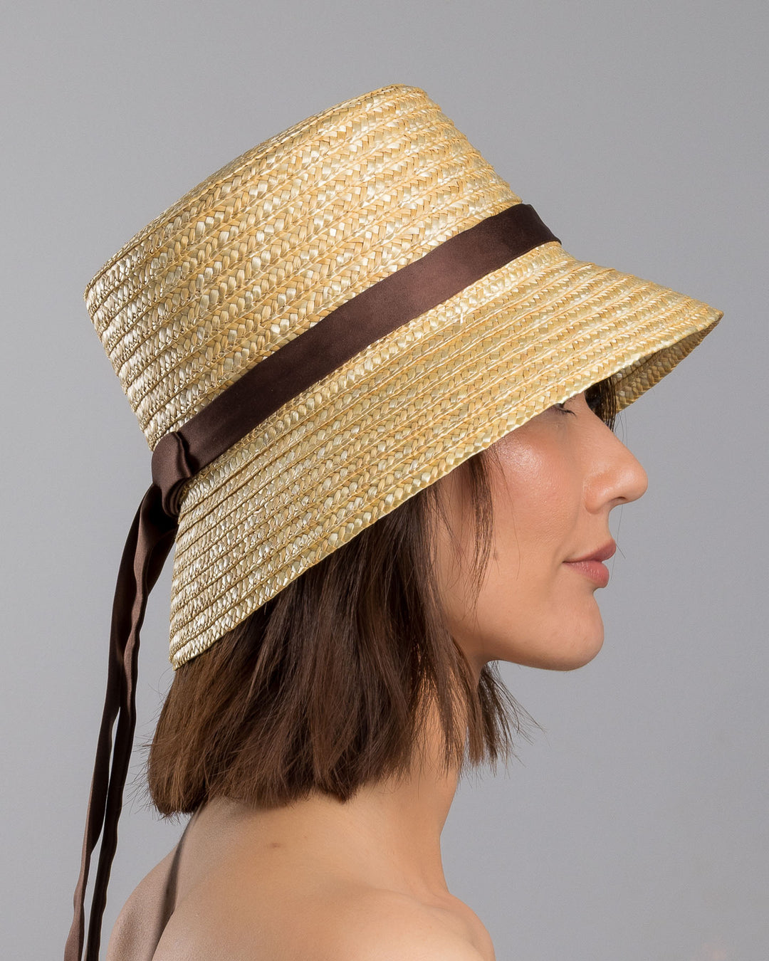 The model is showcasing the right side close up of the natural woven straw bucket hat with a softly sloping brim, finished with a brown ribbon band and long trailing ties at the side. Designed and Made by Merve Bayindir millinery. Designed and made by Merve Bayindir millinery.