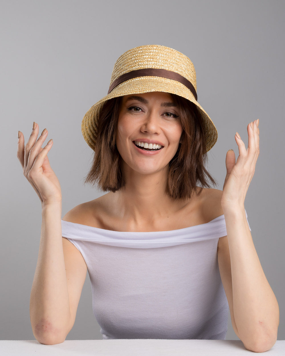 The female model is posing, wearing Brunel, the natural woven straw bucket hat with a softly sloping brim, finished with a brown ribbon band and long trailing ties at the side. Designed and Made by Merve Bayindir millinery. Designed and made by Merve Bayindir millinery.