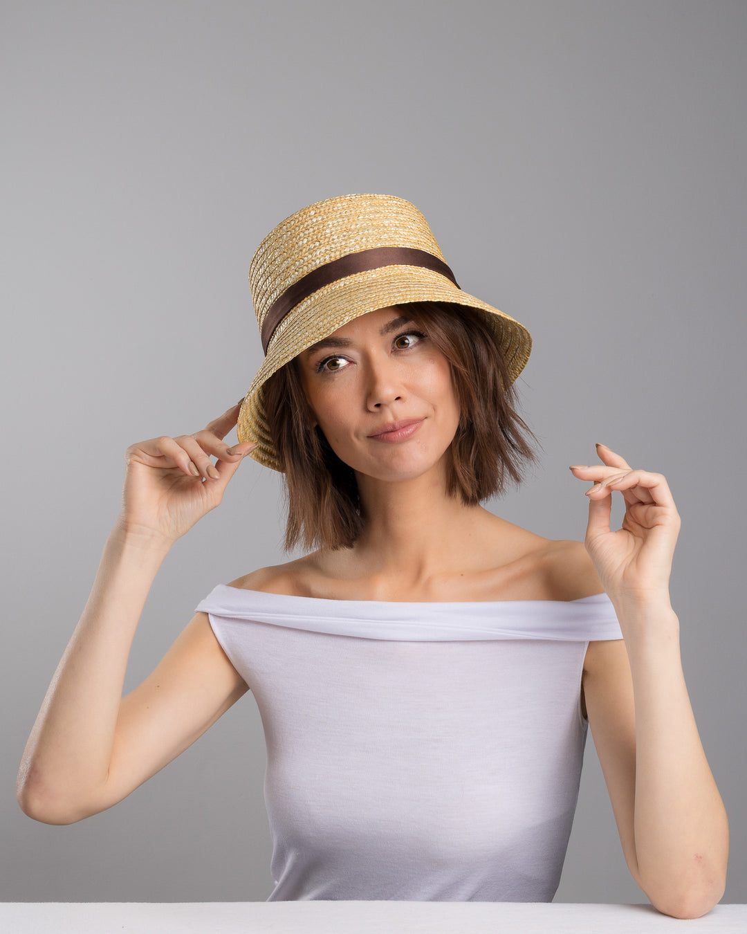The female model is posing, wearing Brunel, the natural woven straw bucket hat with a softly sloping brim, finished with a brown ribbon band and long trailing ties at the side. Designed and Made by Merve Bayindir millinery. Designed and made by Merve Bayindir millinery.