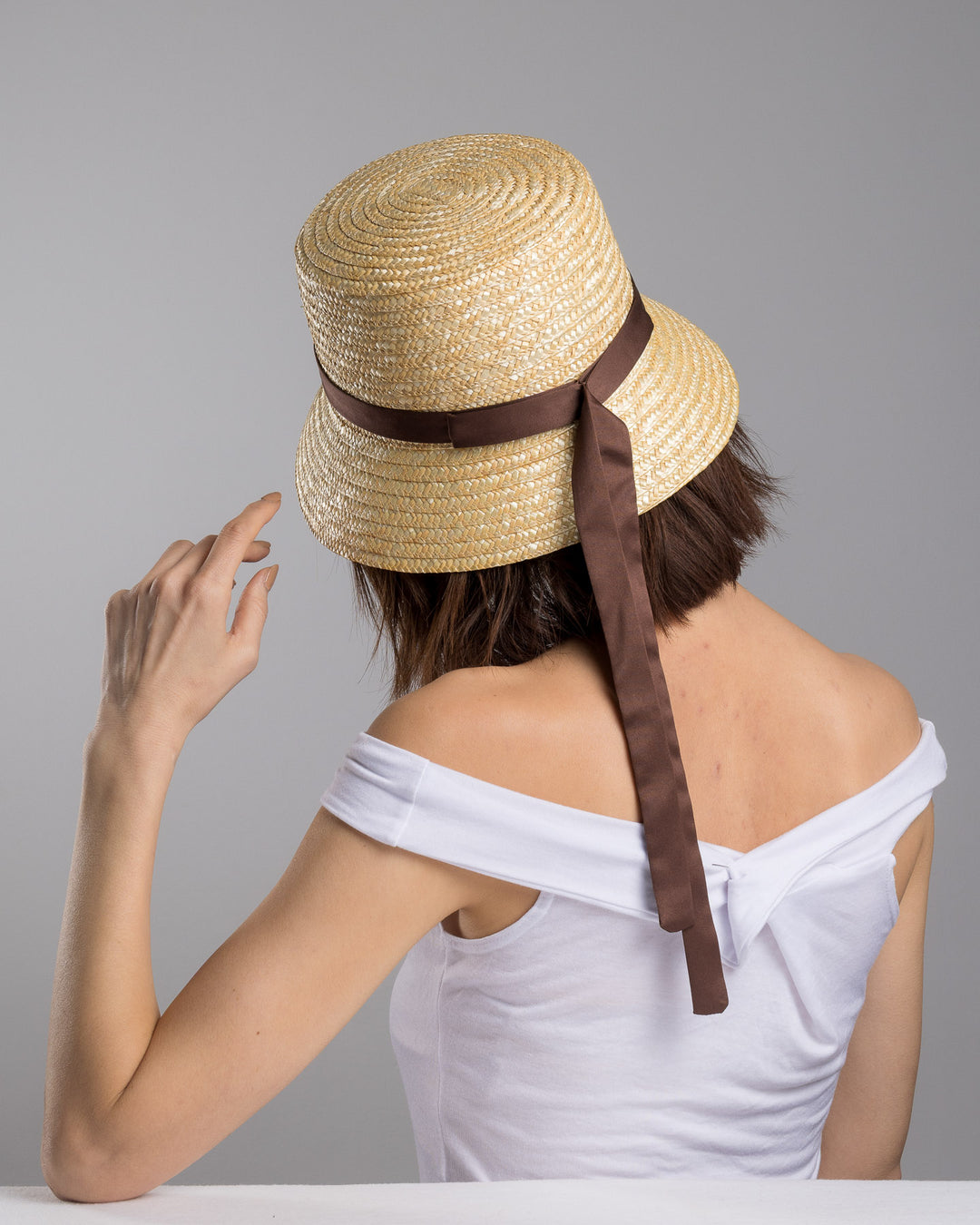 The female model is posing, wearing Brunel, the natural woven straw bucket hat with a softly sloping brim, finished with a brown ribbon band and long trailing ties at the side. Designed and Made by Merve Bayindir millinery. Designed and made by Merve Bayindir millinery.