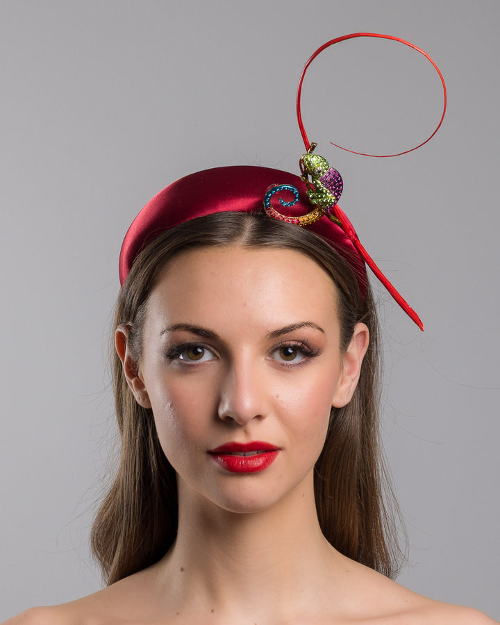 The model is showcasing the front side close up of the red satin padded headband featuring a colorful jeweled chameleon embellishment with a curled tail and a thin red sculptural wire detail extending upward.