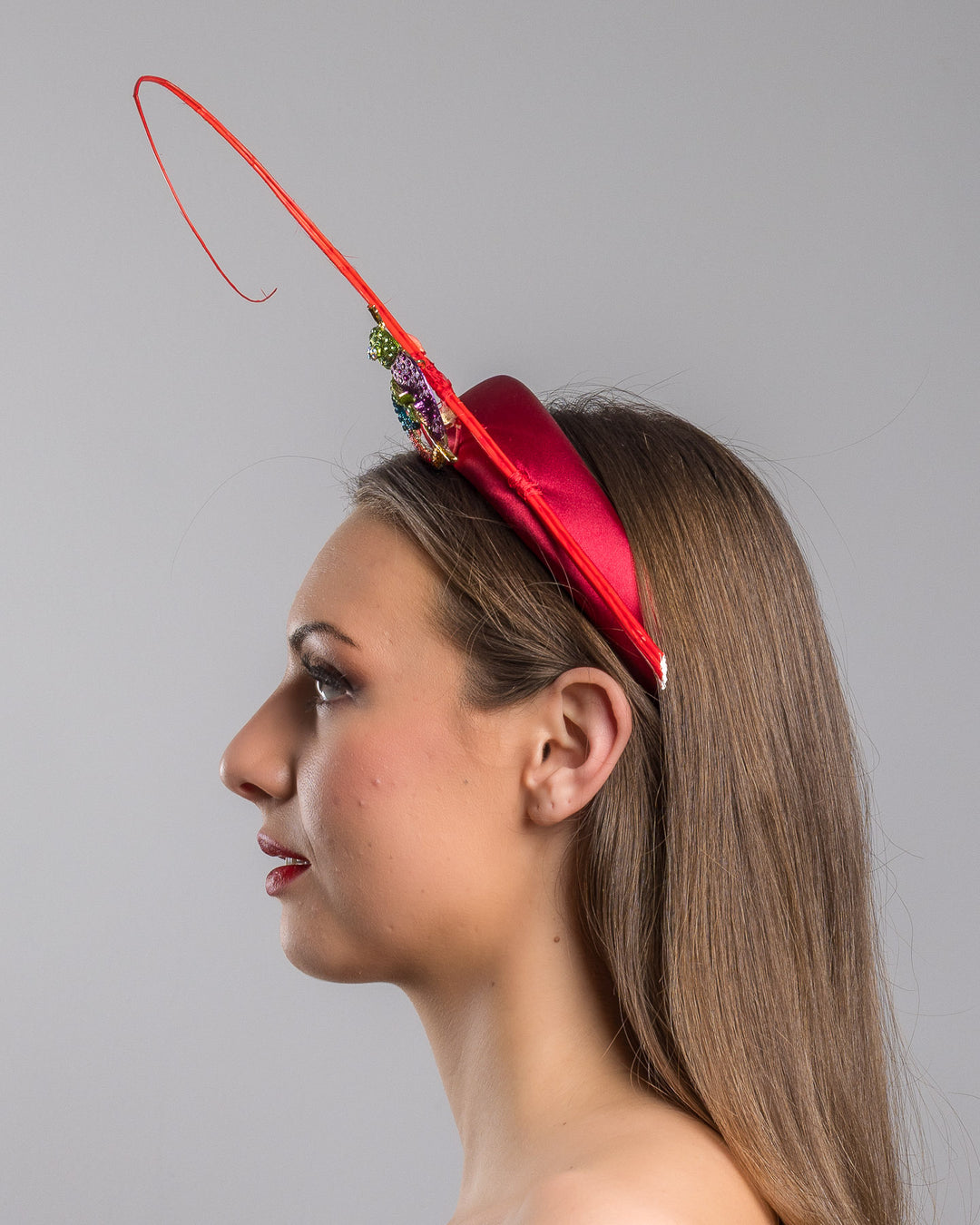 The model is showcasing the left side close up of the red satin padded headband featuring a colorful jeweled chameleon embellishment with a curled tail and a thin red sculptural wire detail extending upward.