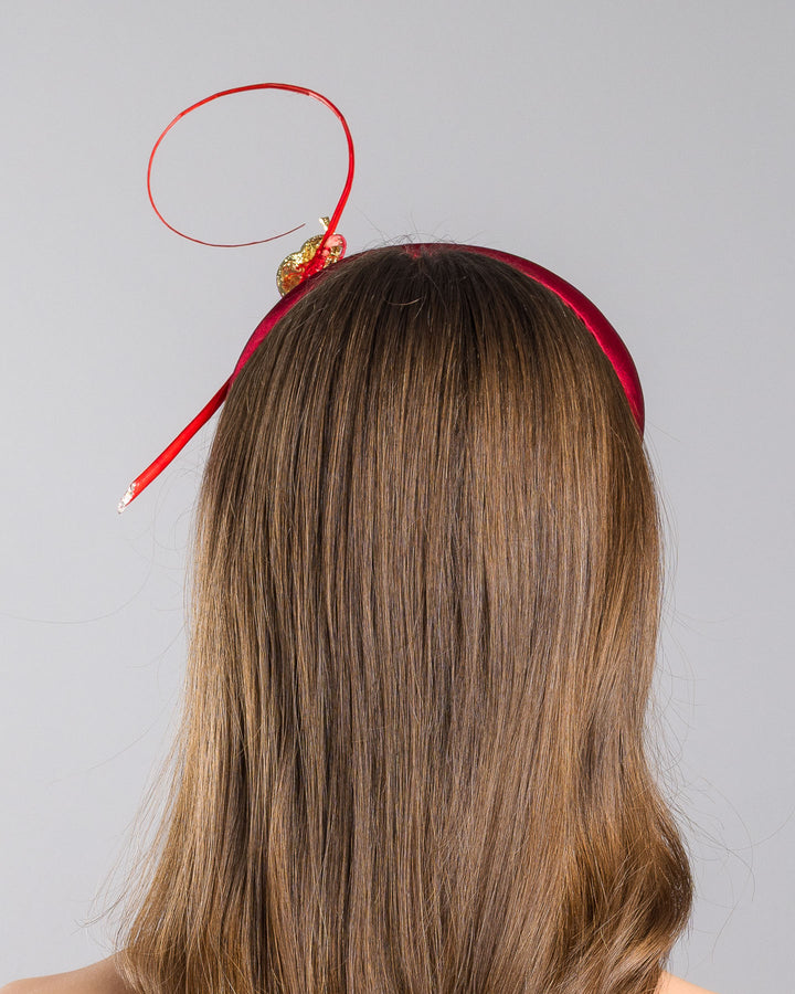 The model is showcasing the back side close up of the red satin padded headband featuring a colorful jeweled chameleon embellishment with a curled tail and a thin red sculptural wire detail extending upward.