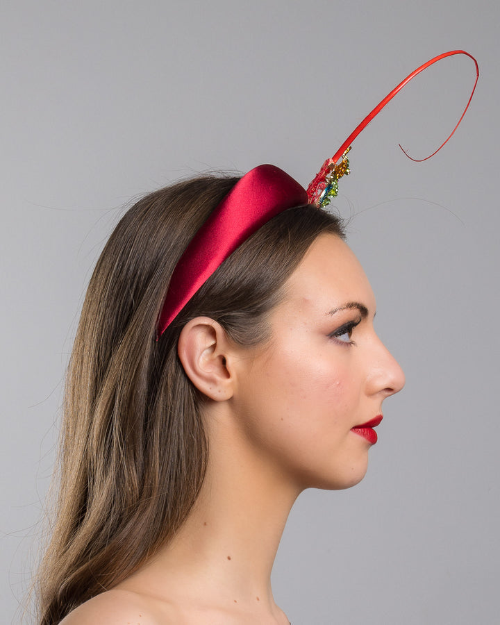 The model is showcasing the right side close up of the red satin padded headband featuring a colorful jeweled chameleon embellishment with a curled tail and a thin red sculptural wire detail extending upward.